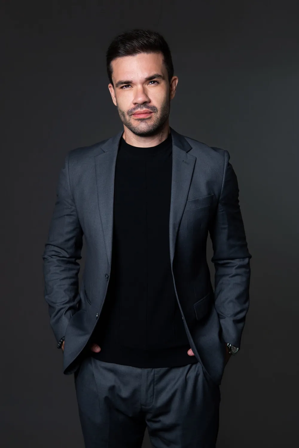 Beto Quintão em pé, de terno cinza e camiseta preta, em retrato de estúdio
