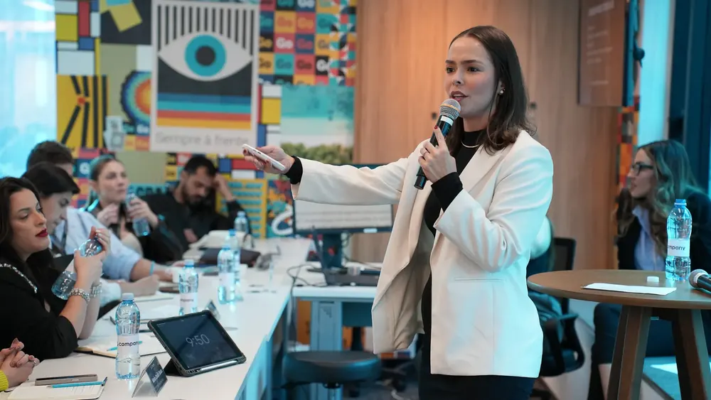Mentora Caroline Mota gesticulando ao microfone para membros sentados em mesas com notebooks